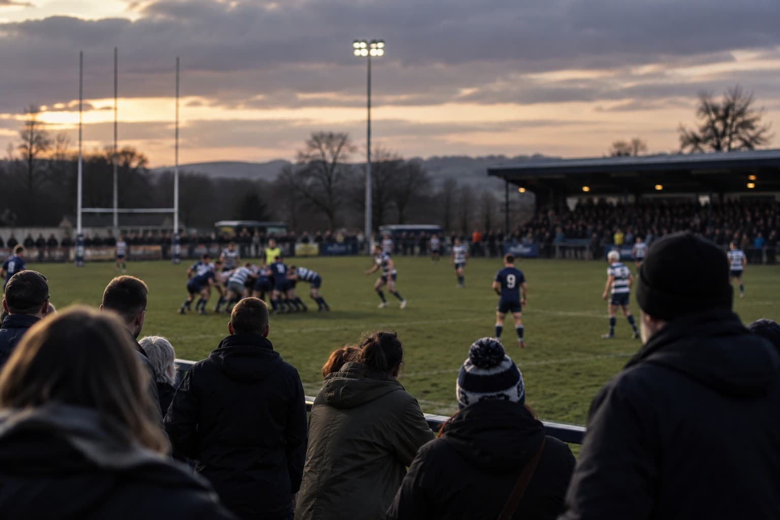 A rugby match at dusk seen over the shoulders of supporters at the rail